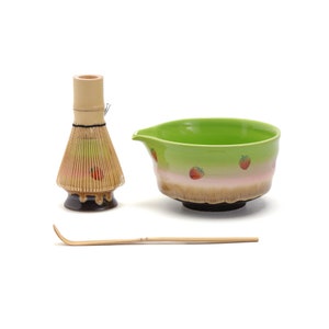 Op de afbeelding: Een matcha theeset met een groene en roze keramische kom met aardbeienmotieven, een bamboe klopper en een bamboe lepel. De kom heeft een schenktuit. De klopper en lepel zijn gemaakt van natuurlijk bamboe.