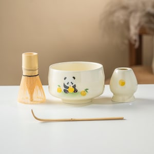 May include: A matcha tea set comprising a cream-coloured ceramic bowl with a panda illustration, a bamboo whisk, a small ceramic container, and a bamboo scoop. The panda is surrounded by yellow fruit illustrations.