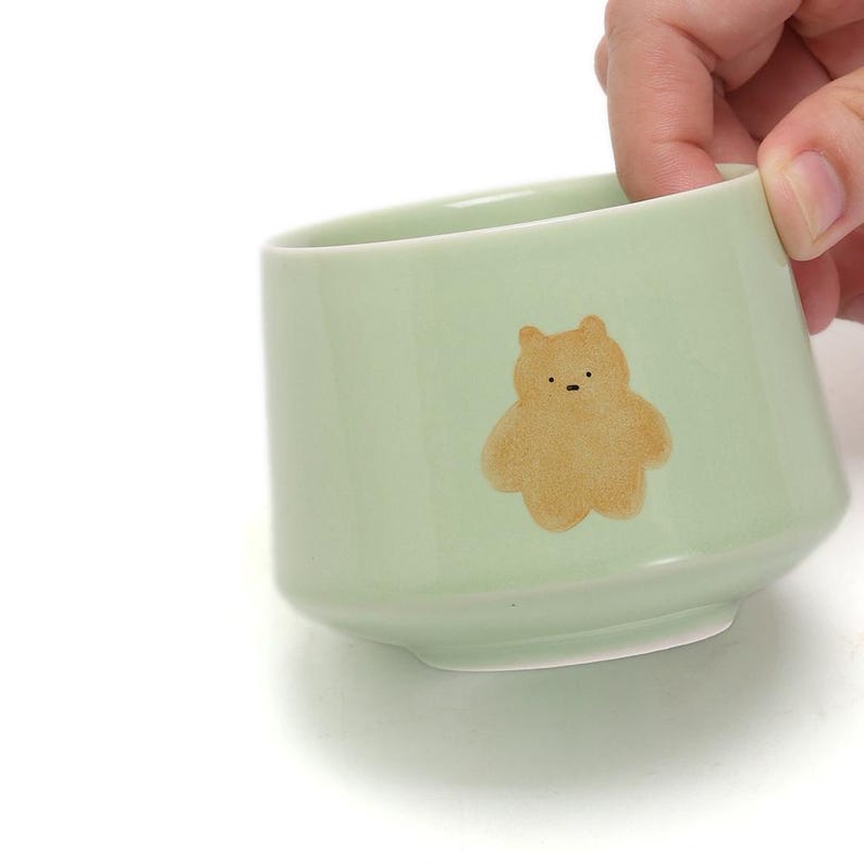 May include: A light green ceramic mug with a brown bear illustration. The mug has a rounded bottom and a wide opening.