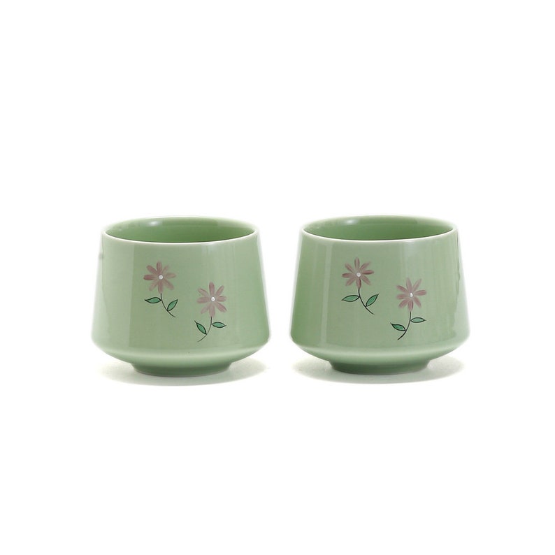 May include: Two green ceramic teacups with a floral design. The cups have a wide base and a flared lip. The flowers are pink and green.