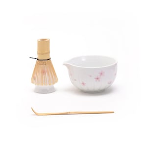 Op de afbeelding: Een witte keramische matcha-kom met een roze bloemenpatroon, een bamboe matcha-garde en een bamboe matcha-schep.
