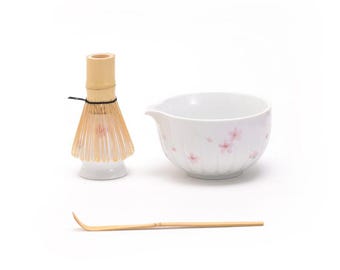 May include: A white ceramic matcha bowl with a pink floral design, a bamboo matcha whisk, and a bamboo matcha scoop.