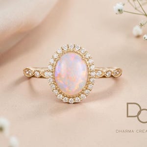 May include: A gold ring with an oval opal gemstone, surrounded by a halo of small diamonds. The ring's band features delicate detailing. The background is a soft, neutral color, with a touch of floral accents.