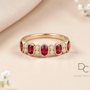 Peut inclure: Une bague en or ornée d'une série de rubis rouges taille ovale alternant avec de petits diamants blancs taille ronde sertis en chaton. L'anneau présente une finition polie et le texte DC DHARMA CREATIONS est imprimé dans le coin inférieur droit.