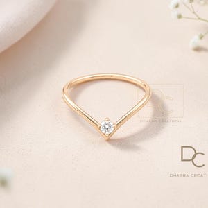 May include: A gold V-shaped ring featuring a single round brilliant-cut white diamond set at the center point. The band is thin and polished, showcasing a minimalist chevron design. The logo for Dharma Creations is visible in the lower right corner.
