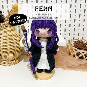 May include: A crochet doll of the character Fern with purple hair, a white dress, and a black coat. The doll holds a staff wrapped in purple yarn with a red bead. Text on the image reads FERN INSPIRED BY SOUSOU NO FRIEREN and PDF PATTERN.
