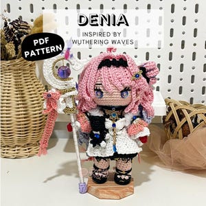 May include: A crochet doll with pink hair, wearing a white and black outfit with blue accents, holding a white and gold staff with a purple bead. The text reads DENIA INSPIRED BY WUTHERING WAVES PDF PATTERN. The doll stands on a wooden base.