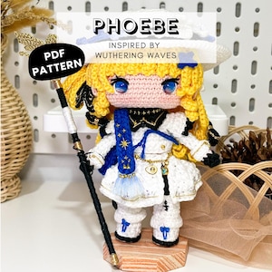 May include: A crochet doll with yellow hair, blue eyes, and a white outfit featuring blue accents, a blue braided accessory, and a black and gold staff. The text PHOEBE INSPIRED BY WUTHERING WAVES and PDF PATTERN is visible on the image overlay.