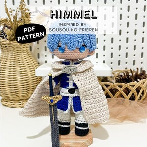 Puede incluir: Una muñeca de ganchillo de un personaje con cabello azul claro y ojos azules, vistiendo un traje azul y blanco con una capa beige. La muñeca sostiene una espada gris oscuro con empuñadura dorada. Texto: HIMMEL INSPIRED BY SOUSOU NO FRIEREN PDF PATTERN.