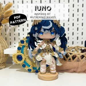 Könnte beinhalten: Handgefertigte Häkelpuppe mit blauen Haaren und einem weißen und goldenen Outfit. Die Puppe steht auf einem Holzsockel. Der Text "JUNO INSPIRED BY WUTHERING WAVES" ist sichtbar. Ein "PDF PATTERN"-Etikett ist ebenfalls vorhanden.