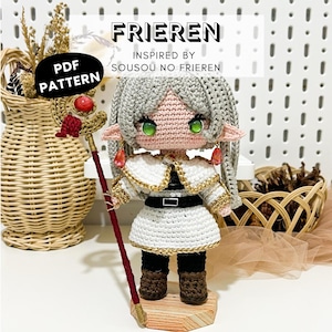 May include: A crochet doll of a character with silver hair, pointed ears, and green eyes, wearing a white tunic with gold trim, a black belt, and brown boots. The doll holds a staff with a red gem and has text reading FRIEREN PDF PATTERN.