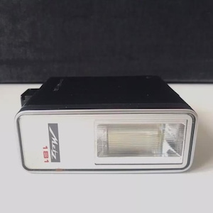 Peut inclure: Un flash d'appareil photo Metz 181 noir avec une bordure argentée. Le flash est de forme rectangulaire avec un panneau avant blanc. Le logo Metz et "181" sont imprimés sur le devant. Le flash est conçu pour les appareils photo.