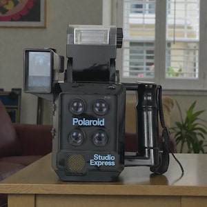 Peut inclure: Appareil photo instantané Polaroid Studio Express noir avec flash intégré et poignée. Le logo Polaroid et l'inscription "Studio Express" sont imprimés sur le devant. Il est posé sur une table en bois.