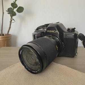 Könnte beinhalten: Schwarze Yashica 108 Kamera mit Zoomobjektiv. Das Objektiv ist mit MC Zoom 28-80mm 1:3.9-4.9 beschriftet. Die Kamera steht auf einer hellbraunen Oberfläche. Im Hintergrund ist eine Pflanze zu sehen.