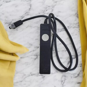 Op de afbeelding: Zwarte Canon afstandsbediening met een zilverkleurige knop en een opgerolde zwarte kabel. De afstandsbediening heeft een zilverkleurige connector. De tekst "Canon" en "Remote Switch" is zichtbaar.