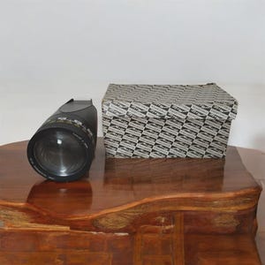 Peut inclure: Un objectif d'appareil photo noir avec des marquages de mesure et un grand élément frontal. Une boîte à motifs avec le texte "mecavox" est à côté de l'objectif. Les articles sont sur une surface en bois poli.