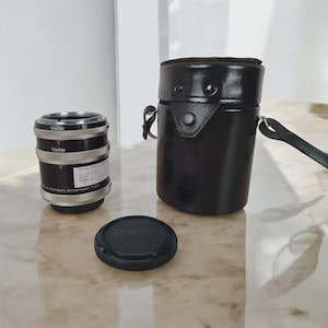 Könnte beinhalten: Ein schwarzes und silbernes Vivitar Automatic Extension Tube für eine Kamera, eine schwarze Canon Objektivkappe und eine schwarze Ledertasche mit Riemen. Auf dem Verlängerungsrohr ist der Markenname Vivitar aufgedruckt. Die Tasche ist zylindrisch.