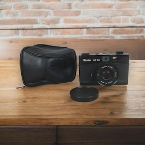 Op de afbeelding: Een zwarte Rollei XF 35 camera met een lensdop en een bijpassende zwarte leren etui. De camera heeft de tekst "Rollei XF 35" en "Sonnar 2.3/40" erop gedrukt. De camera en accessoires liggen op een houten oppervlak.