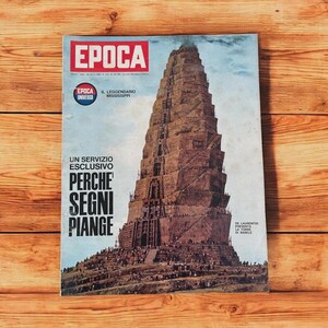 May include: A vintage magazine cover with the title "EPOCA" in red letters. The cover features an illustration of a large, tiered structure, possibly a ziggurat or tower, with the text "PERCHE SEGNI PIANGE". The magazine is on a wooden surface.