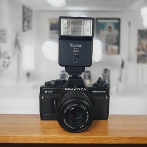 Może przedstawiać: Czarny aparat Praktica BCA z dołączoną lampą błyskową Vivitar Auto 2600. Obiektyw aparatu to Pentacon 1:1.8 f=50mm MC. Aparat stoi na drewnianej powierzchni.