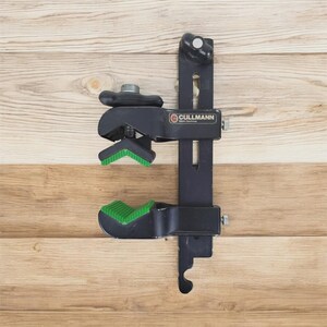 May include: A black Cullmann camera clamp with green rubber grips and a screw-down mechanism. The clamp is mounted on a vertical black bar with a ball head, set against a light brown wooden background.