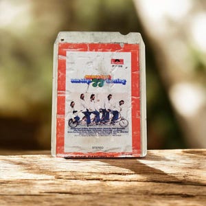 Op de afbeelding: Een vintage 8-track cassette met een wit label en een rode rand. Het label bevat de tekst James Last Non Stop Dancing, een foto van vier personen op een tandemfiets, het Polydor-logo en een lijst met muzieknummers.