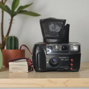 Op de afbeelding: Zwarte Yashica AF-mini camera met een zwarte leren hoes. De camera heeft een rode riem en een stapel kleine witte kaarten. De tekst "YASHICA AF-mini" en "YASHICA LENS" is zichtbaar.