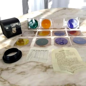 Op de afbeelding: Een verzameling camerafilters in diverse kleuren, waaronder groen, oranje en blauw, gepresenteerd in transparante plastic cases. Een zwart leren etui met de tekst "Scamo 52" en een zwarte lensadapter zijn ook aanwezig. Inclusief gedrukte documentatie.