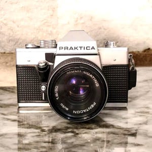 May include: A silver and black Praktica camera with a Pentacon lens. The lens has the words "Multi Coating" and "auto" printed on it. The camera is on a marble surface.