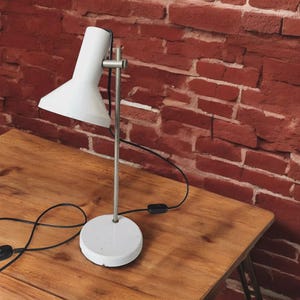 May include: A white desk lamp with a conical shade and a circular base. The lamp has a silver-coloured, adjustable arm and a black power cord. The lamp is on a wooden table in front of a red brick wall.