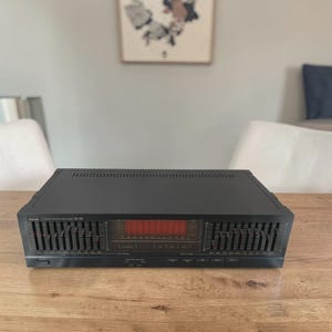 Op de afbeelding: Een zwarte Sansui SE-80 stereo grafische equalizer. Het audio component heeft meerdere schuifregelaars en een rood LED-display. Het apparaat staat op een houten tafel. De achtergrond omvat een schilderij en een witte stoel.