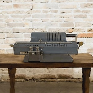 Op de afbeelding: Een vintage grijze mechanische rekenmachine met een handvat en meerdere wijzerplaten. De rekenmachine is gemaakt van metaal en staat op een houten tafel. De achtergrond is een bakstenen muur.