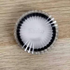 May include: A clear, circular object with a white interior and a black border. The word "Kenko" is printed in the centre. The object has a ridged, transparent outer edge.