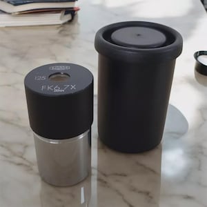 Könnte beinhalten: Zwei zylindrische Komponenten. Eine ist schwarz mit dem Text "OLYMPUS" und "FK6.7X JAPAN" aufgedruckt und hat eine silberne Basis. Die andere ist ein hoher, schwarzer Zylinder mit flachem Deckel. Beide stehen auf einer hellen Oberfläche.