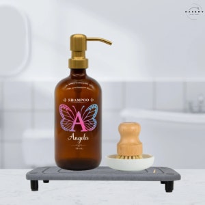 May include: A brown glass amber bottle with a gold pump dispenser. The bottle has a pink and blue butterfly with the letter A and the name Angela printed on it. The bottle is labeled "Shampoo 16 oz.". The bottle is sitting on a gray tray with a wooden brush and a white dish.