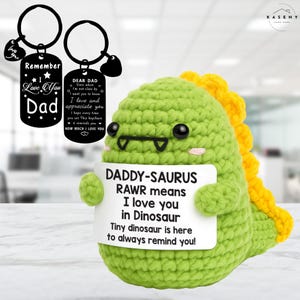 May include: A green crocheted dinosaur holding a sign that reads "DADDY-SAURUS RAWR means I love you in Dinosaur." Two black keychains with engraved messages for "Dad" are also visible. The dinosaur has yellow spines.