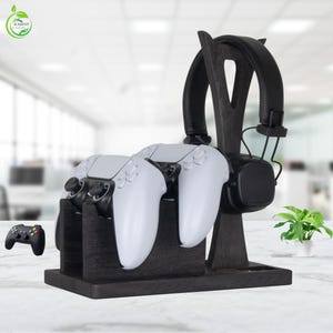 May include: A black wooden gaming organizer stand featuring a vertical support for a pair of black over-ear headphones and two front-facing slots designed to hold two white game controllers. A small black game controller is visible to the left.