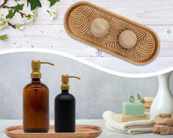 May include: Two amber glass soap dispensers with gold pumps, and a black glass soap dispenser with a gold pump, are displayed on a wooden tray. The tray has a carved design and is oval shaped.