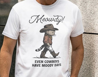 May include: White t-shirt with a cartoon illustration of a cat wearing a cowboy hat and boots. The text "Meowdy!" is above the cat and "Even cowboys have moody days" is below the cat.