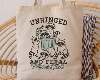 May include: Cream canvas tote bag featuring the text "UNHINGED AND FERAL Moms Club" and a raccoon graphic. The raccoons are depicted around a bin. The bag has two long handles and is on a wicker chair.