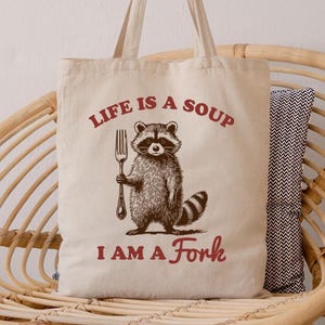 May include: Beige tote bag with the text "LIFE IS A SOUP I AM A Fork" in red. The bag features a raccoon illustration holding a fork. The bag has long handles and is made of a canvas-like material.