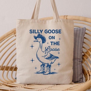 May include: A white canvas tote bag with a blue illustration of a goose wearing a cowboy hat and boots. The text "Silly Goose On The Loose" is printed on the bag.