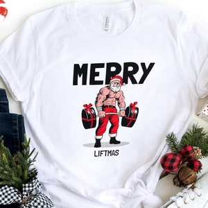 May include: White t-shirt with a graphic of a muscular Santa Claus lifting weights. The text "Merry Liftmas" is printed below the image.