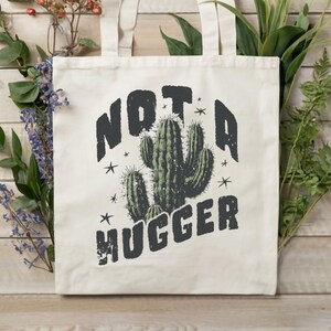 May include: Cream-colored tote bag with the text "NOT A HUGGER" above a cactus illustration. The cactus is green with dark green shading. The bag has two cream-colored handles and is set against a wooden background with flowers.