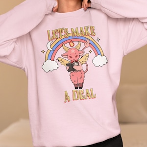 May include: Pink sweatshirt with a cartoon devil holding a book and a rainbow above it. The text "Let's Make A Deal" is printed on the sweatshirt.