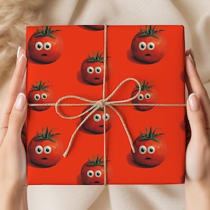 May include: Red gift wrap featuring a repeating pattern of cartoon tomatoes with surprised expressions. The wrap is tied with twine and held by hands with manicured nails. The tomatoes have green stems and white eyes.
