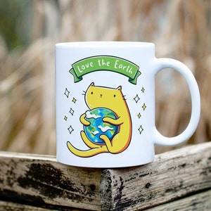 May include: White ceramic mug with a yellow cat holding a blue and green globe. The mug has the text "Love the Earth" in green letters on a white banner.