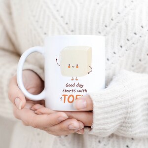 Może przedstawiać: Biały kubek ceramiczny z kresk&oacute;wkowym blokiem tofu z uśmiechającą się twarzą i tekstem "Good day starts with TOFU".