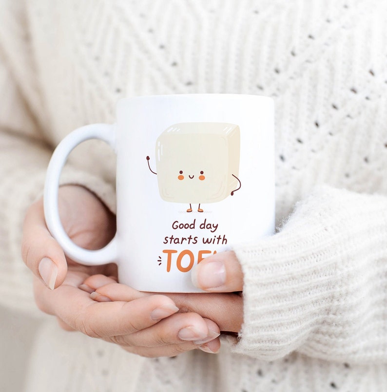 Może przedstawiać: Biały kubek ceramiczny z kresk&oacute;wkowym blokiem tofu z uśmiechającą się twarzą i tekstem "Good day starts with TOFU".