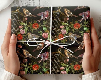 Può includere: Un regalo avvolto con uno sfondo nero adornato da un motivo ripetuto di uccelli, gabbie per uccelli e fiori nei toni del rosso, rosa e verde. Il regalo è legato con un nastro bianco.